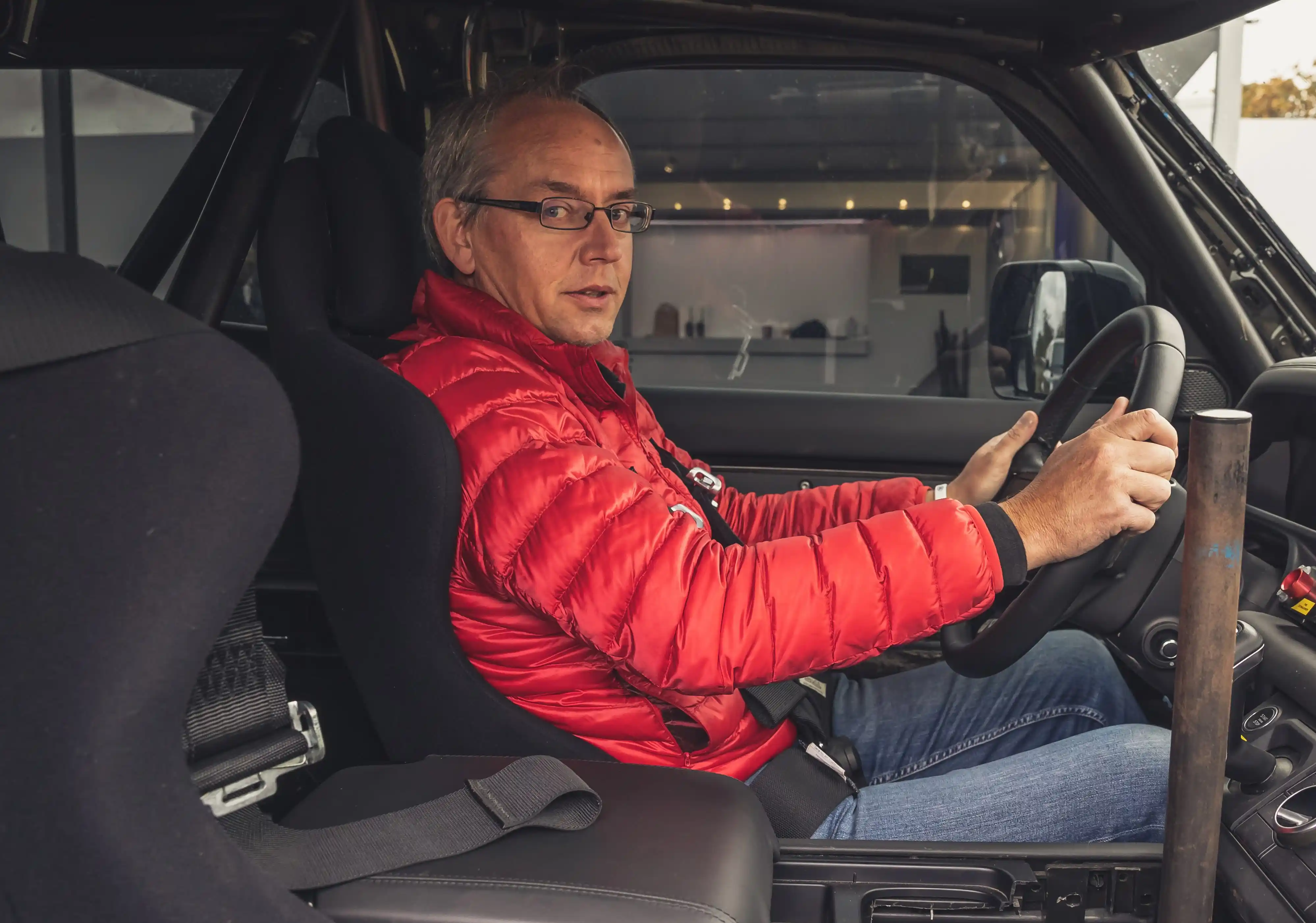Mark Smyth wearing glasses and a red puffer jacket sits in the driver’s seat of a vehicle, holding the steering wheel and looking toward the camera.
